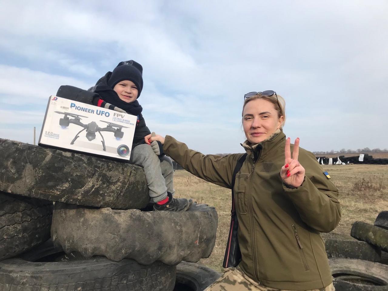 Savva and his mother outdoors beside stacked tires, with Savva holding a boxed toy drone